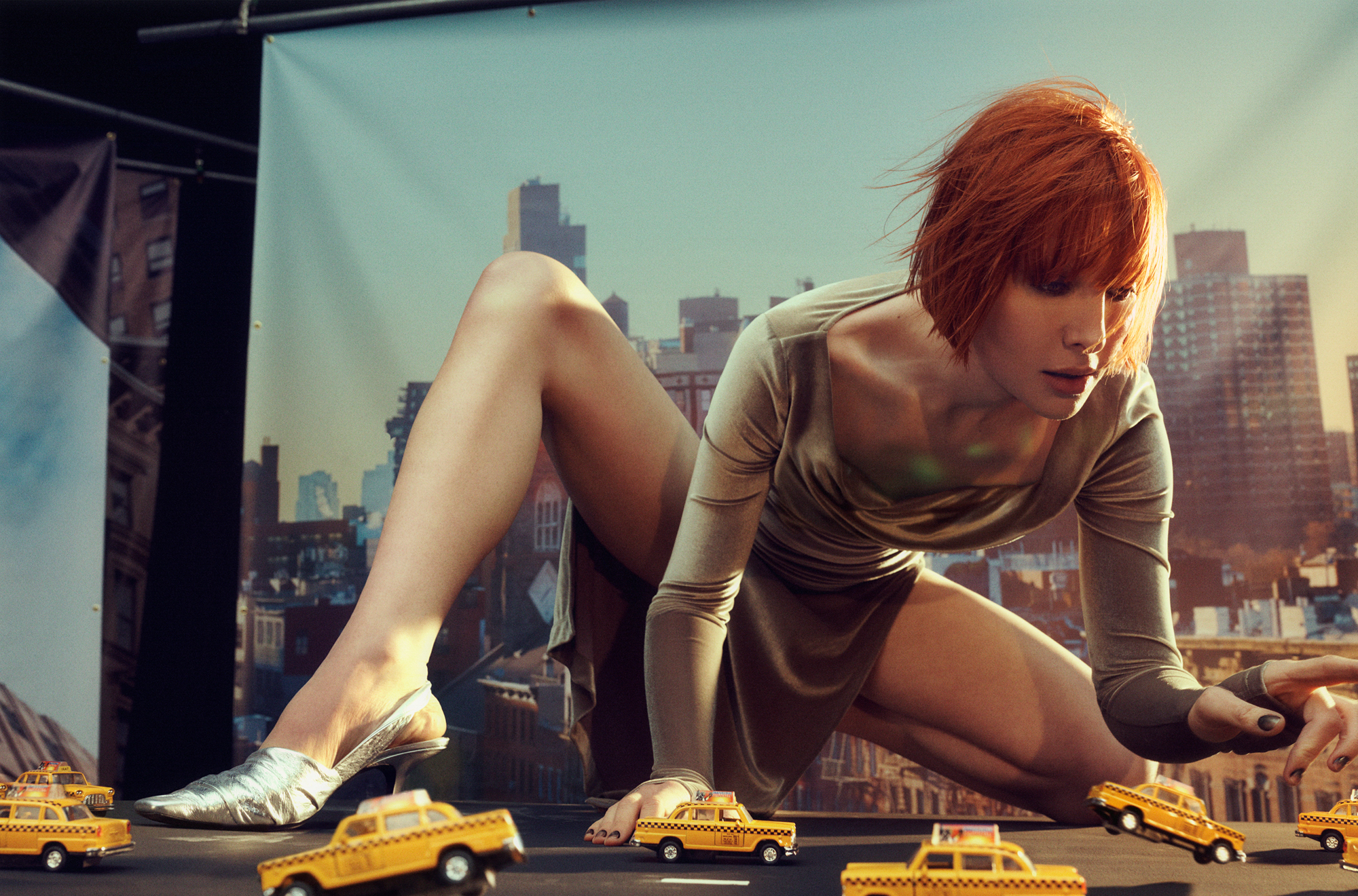 Hari Nef is knelt down on the ground with one leg slightly up, she is looking down at small miniature taxi cab cars. Backdrops of the New York City skyline are visible in the background, with the edges of the backdrops and the support poles visible. The image has a feel as if she is a giant looking down at the taxi cabs below. 