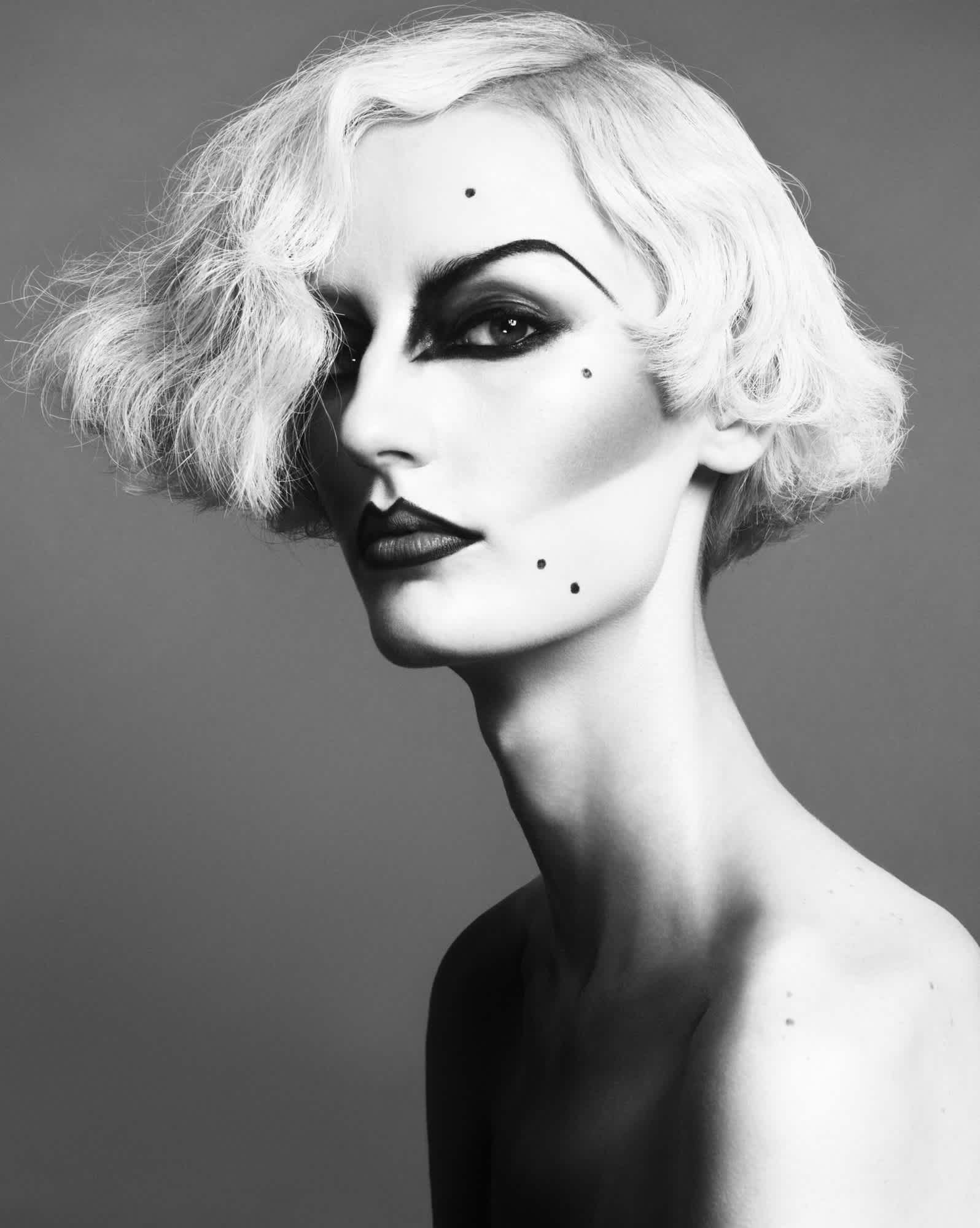 Model is in a partial profile pose. They have a blonde bob haircut, heavily contoured cheeks, deep eyeshadow and brows, and moles applied with makeup. The model is only visible from the shoulders up.