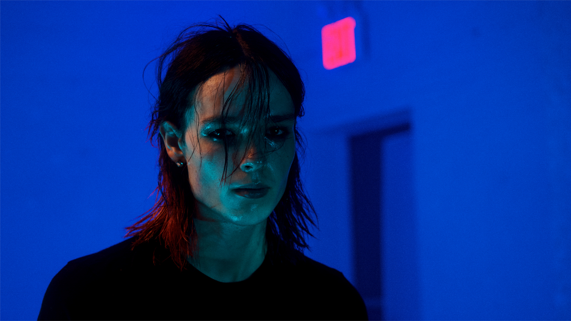 A male model is standing in front of a blue / purple toned background. A doorway is visible to the right. He has blue toned light shining on the face from below. The face is wet and shiny, and he has messy shoulder length hair. 