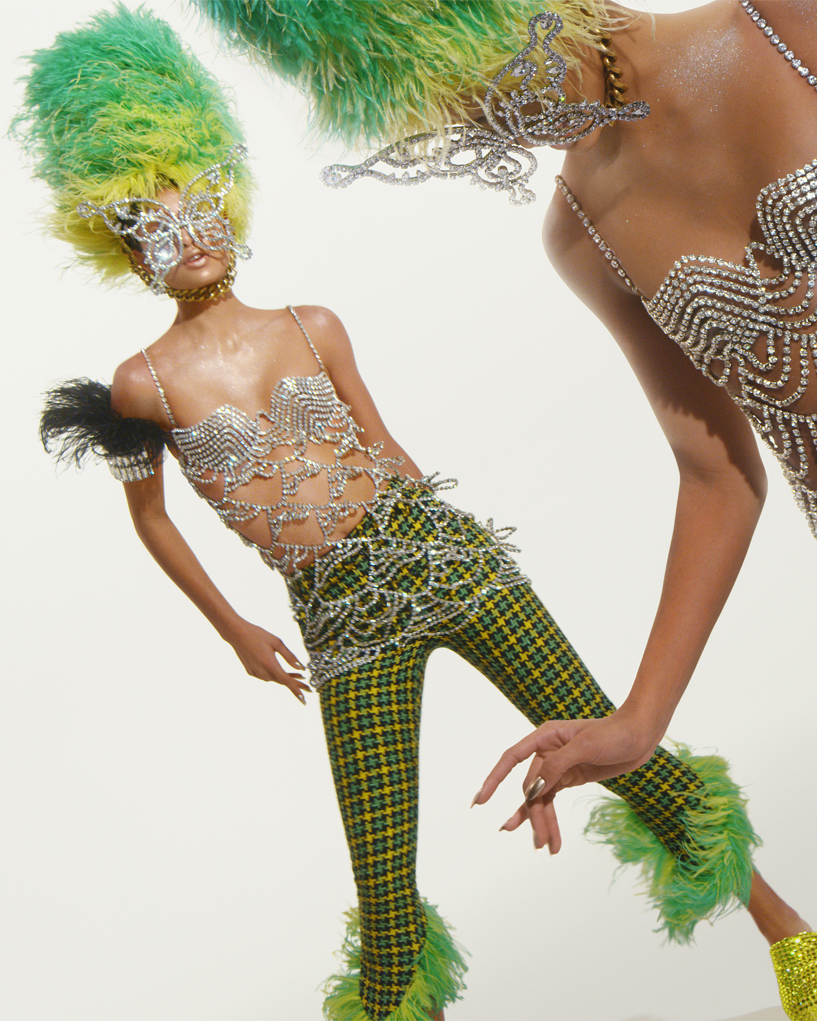 Two models are within the frame at a titled angle. They are both wearing yellow and green checkered pants, bra with jewels, jeweled face covering, and a feathery green and yellow hat. 