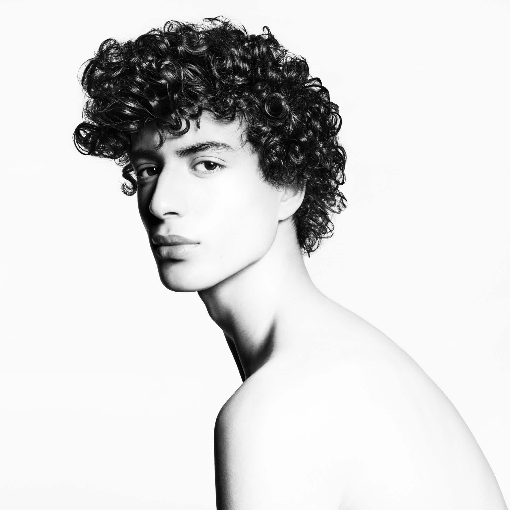 Black and white image of a male model with curly dark hair. His body is positioned to the side, and he is turning to look directly at the camera.