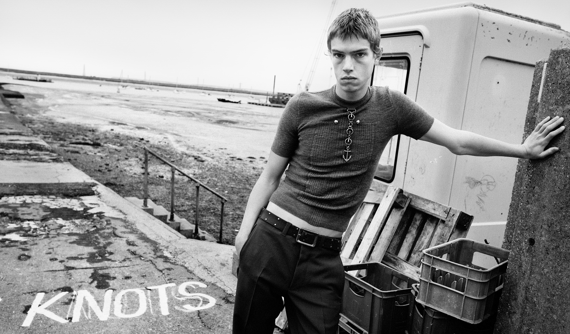 A man is standing with his arm resting on a column in the right hand corner of the image. He is looking directly at the camera and wearing a fitted short sleeved shirt and pants. Behind him is wet marsh lands where there is a staircase that goes down onto the shore. 