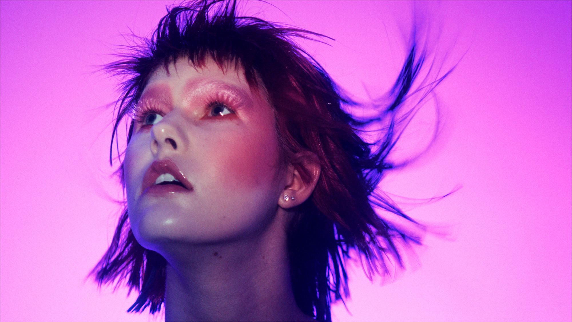A woman in a choppy bob is in the frame in front of a pink background. She has pink blush that is draped around the cheeks and on the temples. Her brows are blocked out with a pink shadow, and she has larger false eyelashes. Her lips have a glossy pink color on them. 