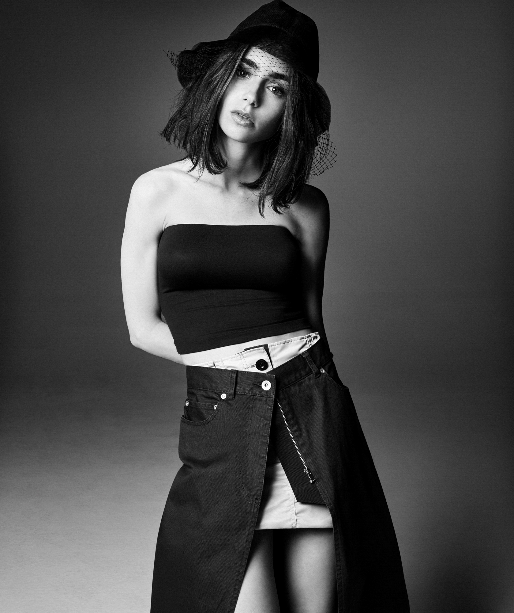 Lily Collins is in a black tube top, layered denim skirt, and a black hat with lace coming out. She has her arms behind her back, head tilted, and looking directly at the camera. 