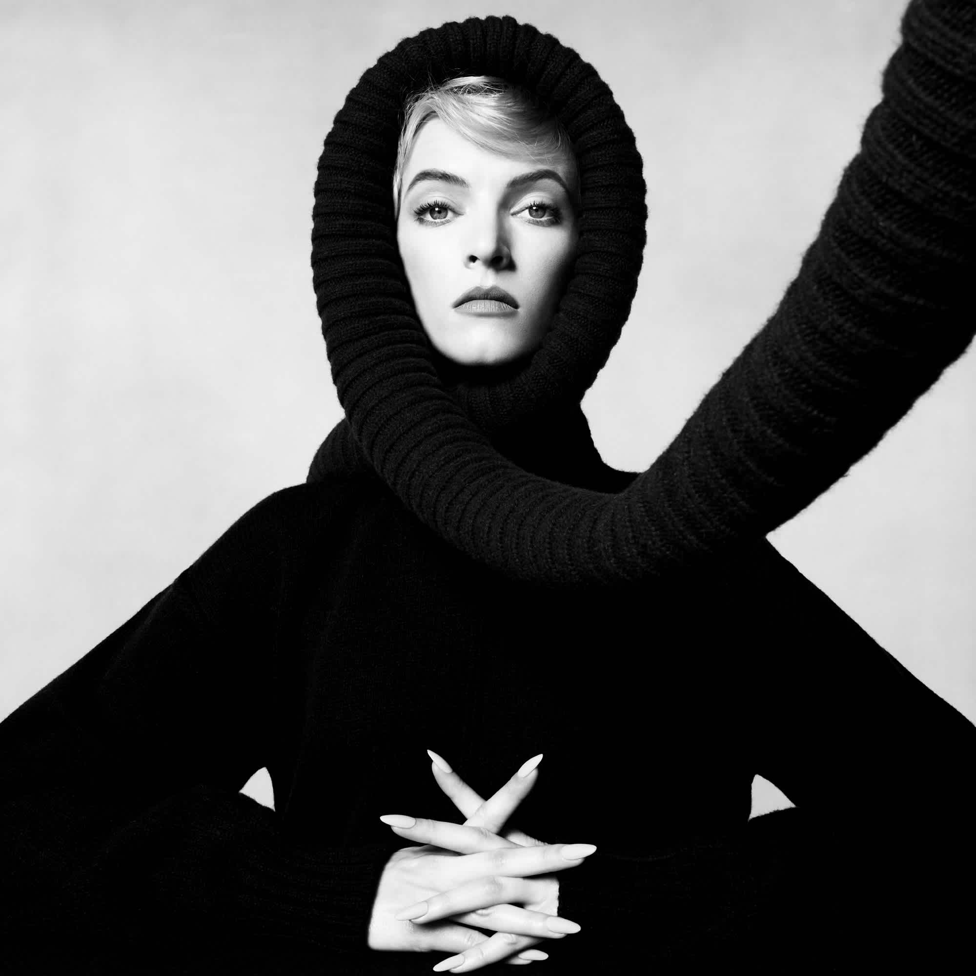 A woman is facing the camera directly wearing a thick black sweater and a scarf over her head that is coming towards the camera. She has her hands clasped together partially.