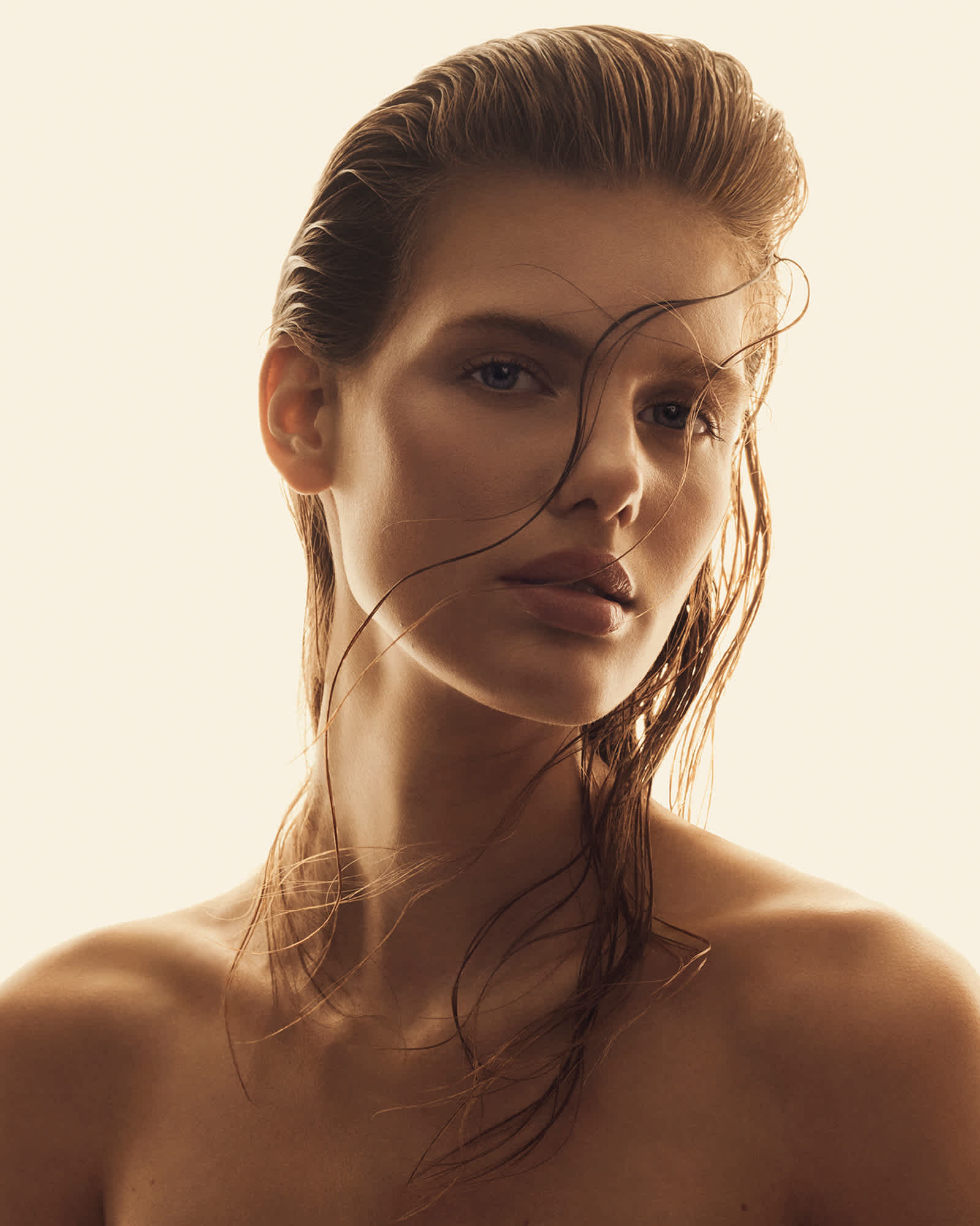 A woman is seen from the shoulders up looking directly at the camera with her head at an angle. Her blonde hair is combed back and small wet stringy pieces of hair are going over her face. She is posing in front of a golden background.