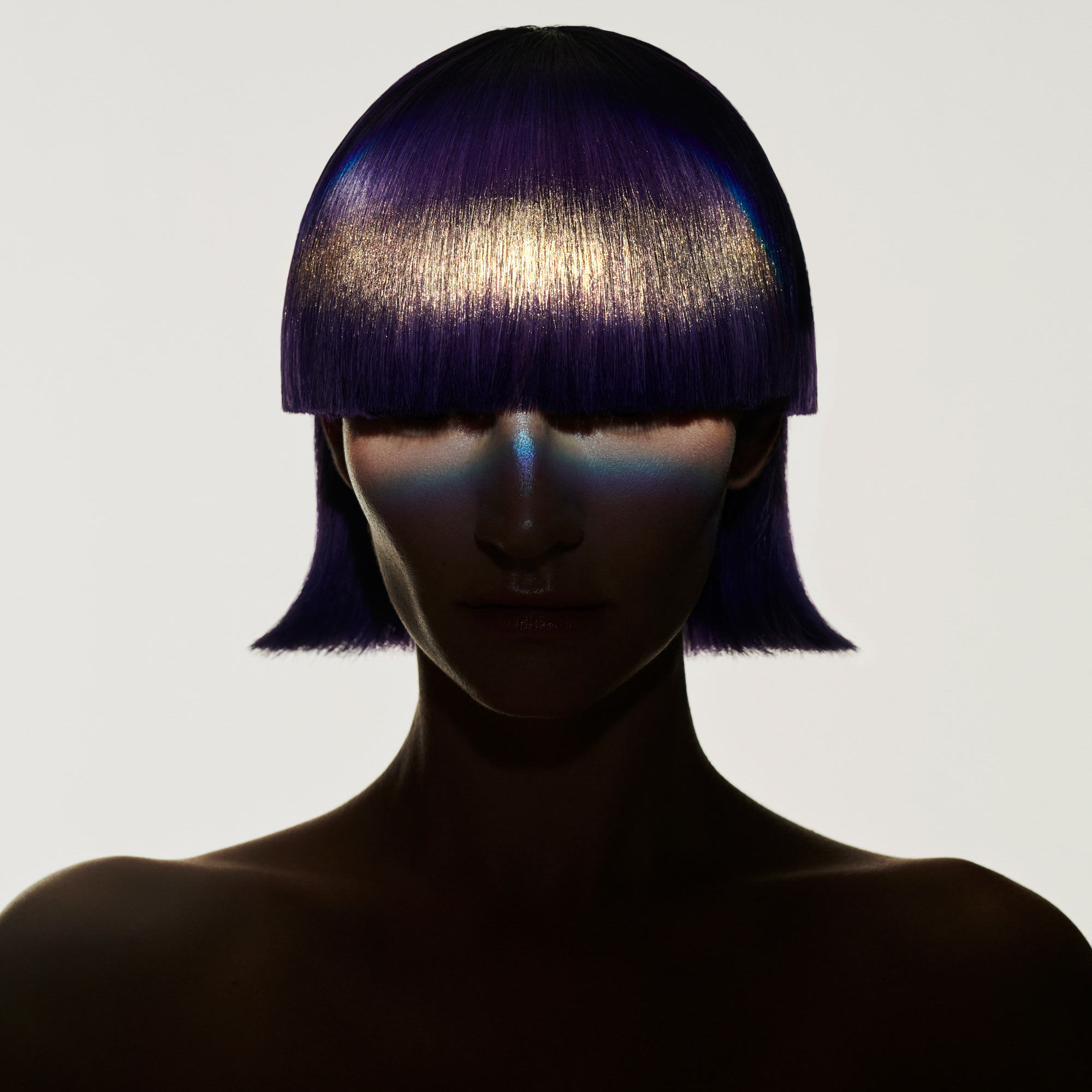 A model is facing the camera with a severe cut fringe and hair that swoops out slightly at the edges near the neck. A spotlight is hitting the model right in the center of their face and it is illuminating the gold makeup in the hair. 