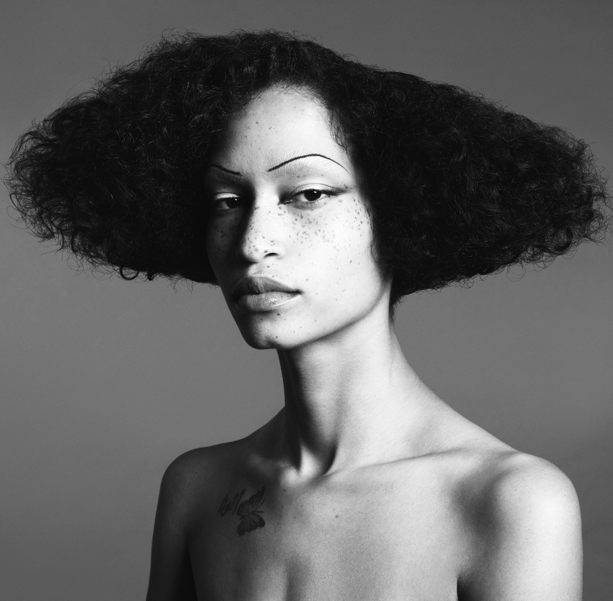 A woman is seen from the shoulders up in the middle of the frame. There is minimal shadow around the lash line of the eyes, and a thin arched brow is highlighted. Their hair is in a sculpted mushroom afro. 