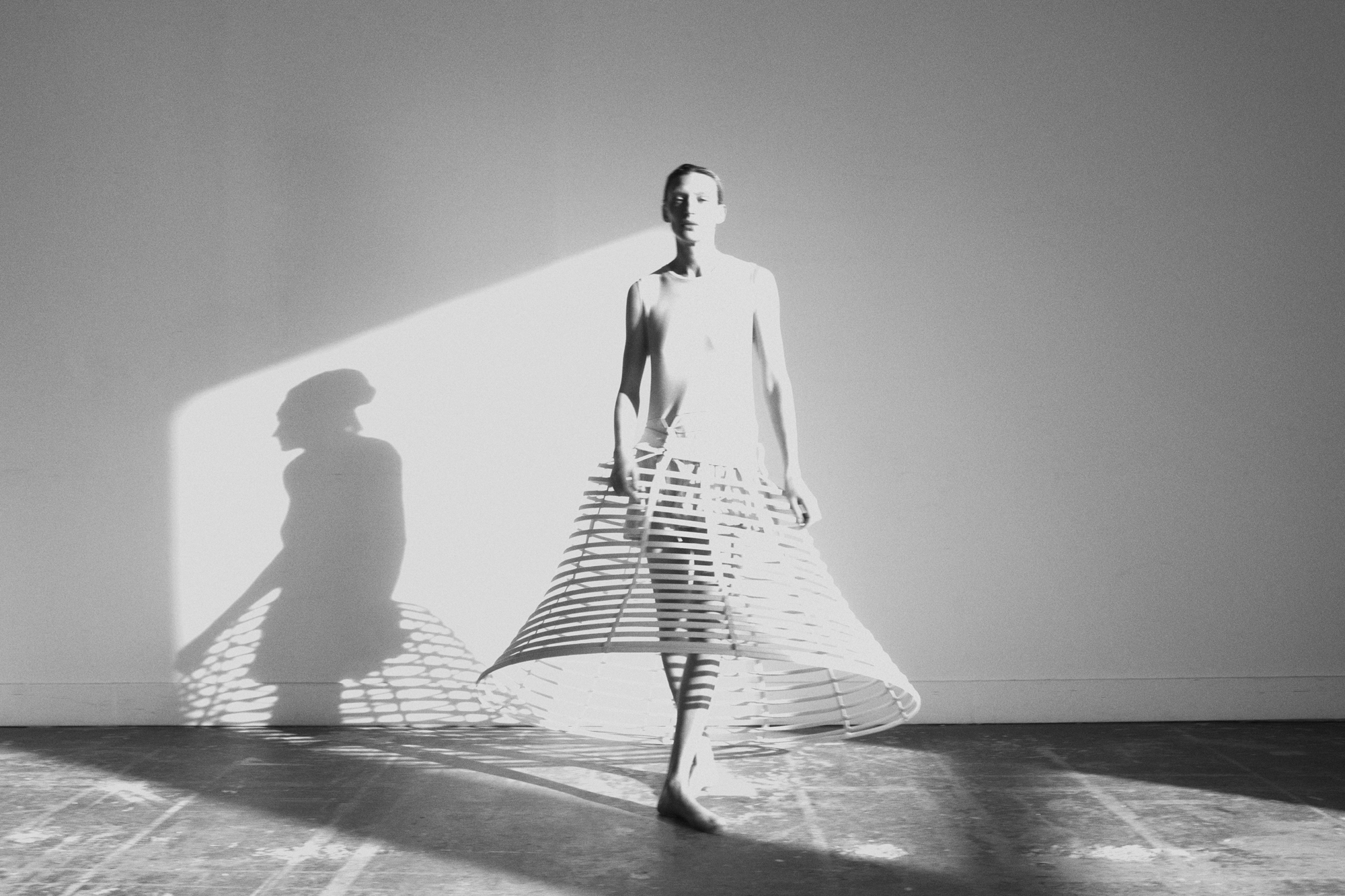 Model is standing in the center of the frame with a structured skirt. The skirt has rectangular openings in it. Model is standing in front of a blank wall, and a sparse concrete floor. 