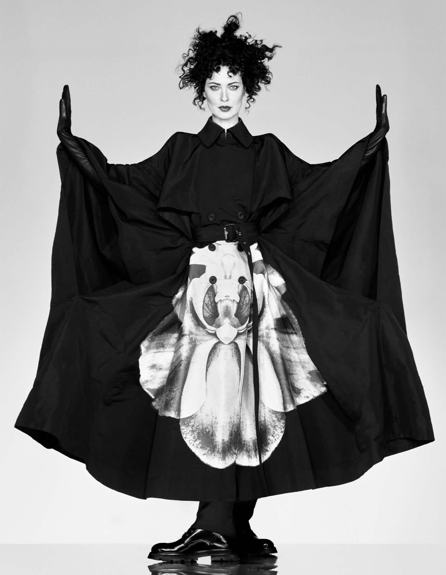 Shalom Harlow is standing in the center of the frame with her arms up. Her hair is very curly, and she is wearing a full length garment that is black with a floral motif in the middle lower half of the garment.