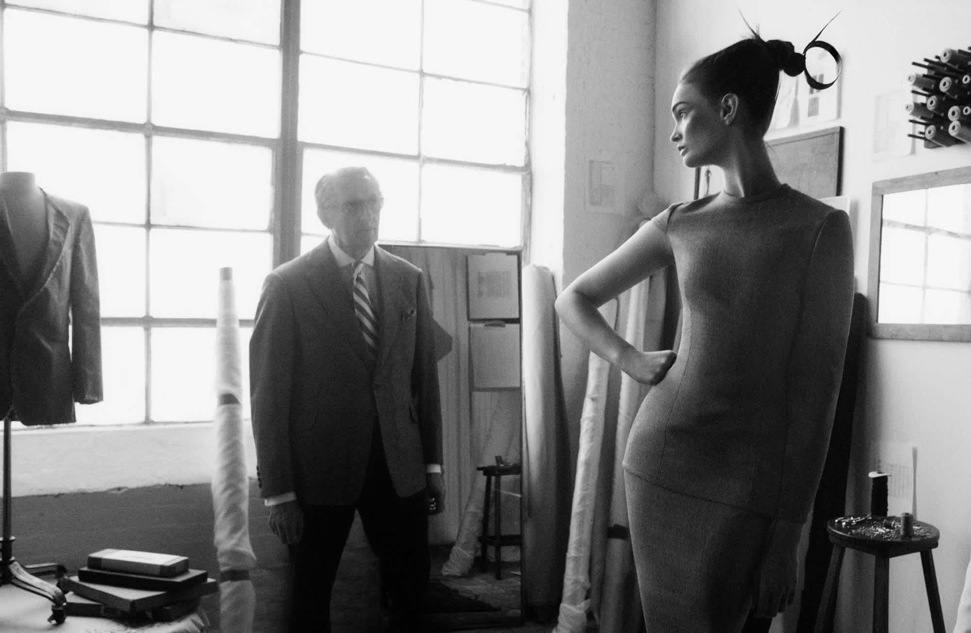 Lulu Tenney has her hand on her hip and is facing a wall of windows. There is an older man that is looking at her. A fitting looks to be going on, and the man is looking at her to analyze the fit of the dress.