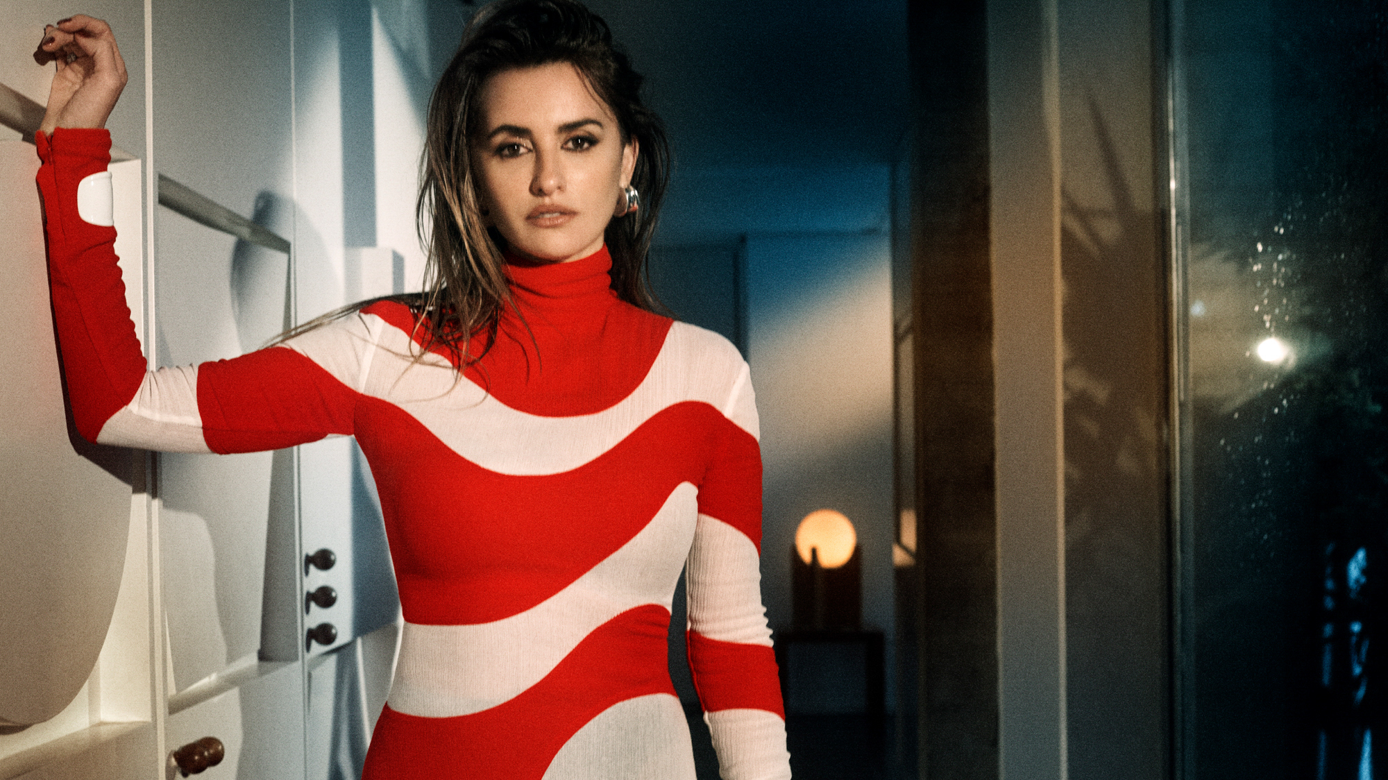 Portrait of Penelope Cruz within a more residential interior. She has her arm resisting on the wall next to her. She is wearing a red dress with white swirling lines that are apart of the design. 