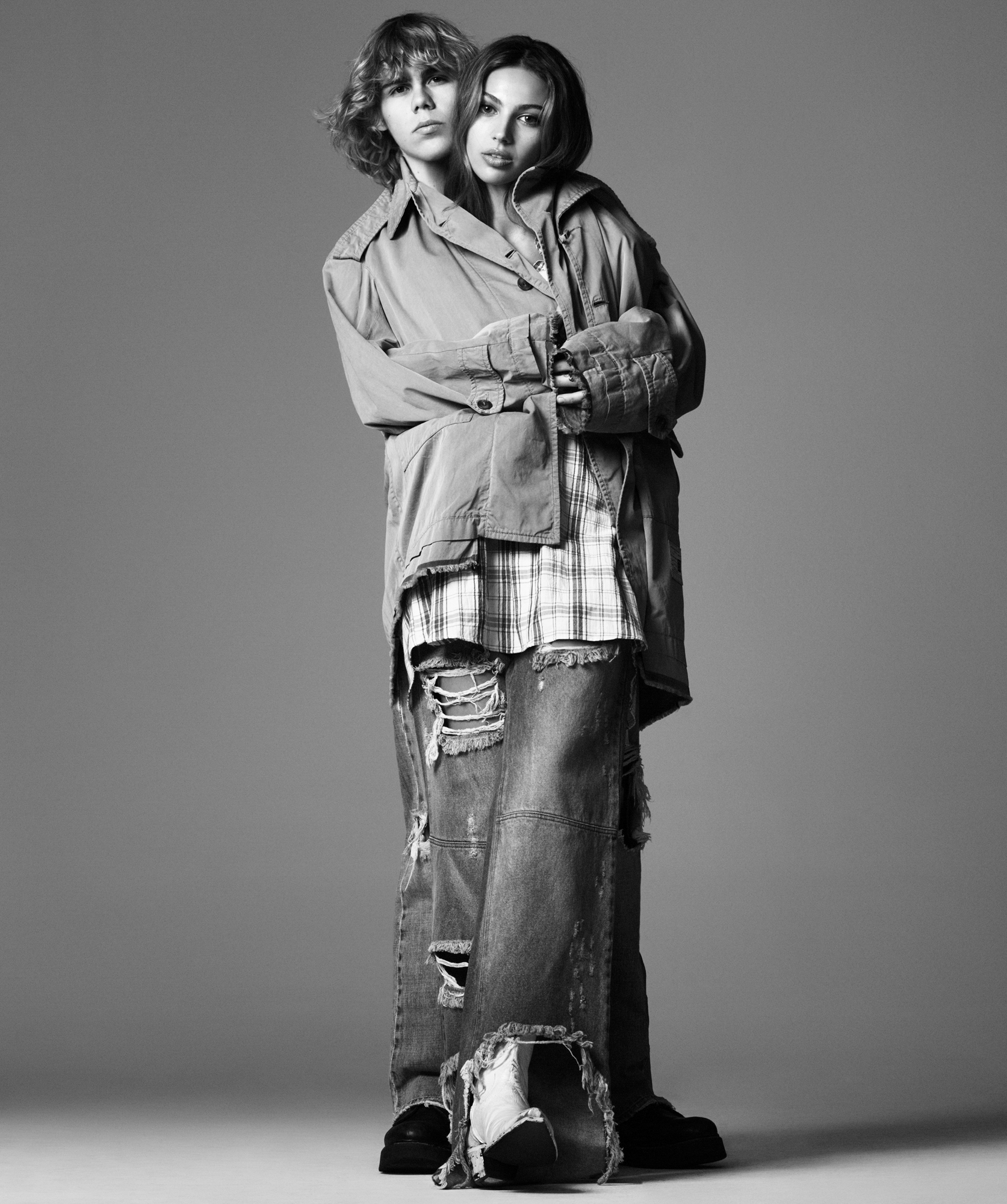 Kid Laroi and a woman model are embracing within a large coat. Kid Laroi is behind her, and both are looking directly at the camera. They both have grungy plaid shirts on and ripped jeans. 