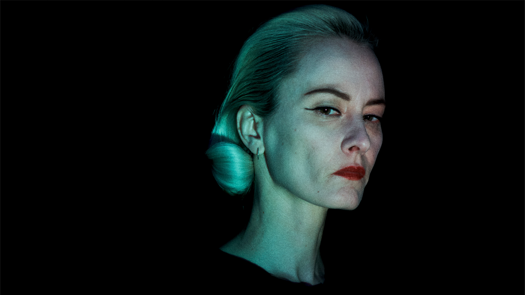 A model against a black background has her blonde hair in a coiffed hairstyle with hair curled inward near the upper neck. The model has a small black winged liner, and a red lip. She is illuminated in a blue toned light. 