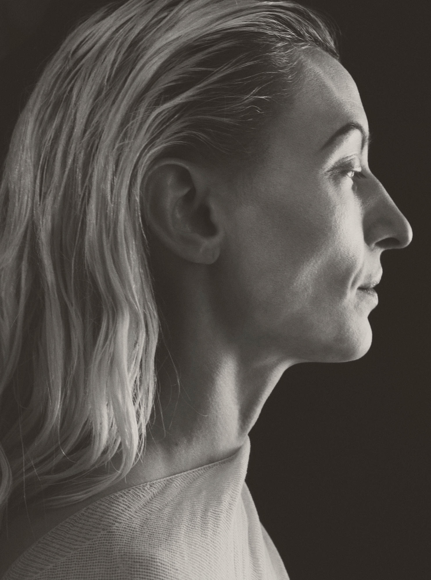 A woman is positioned in profile. She has slicked back hair that is blonde. Light is hitting the model from the front of her face, the background is a solid black. 