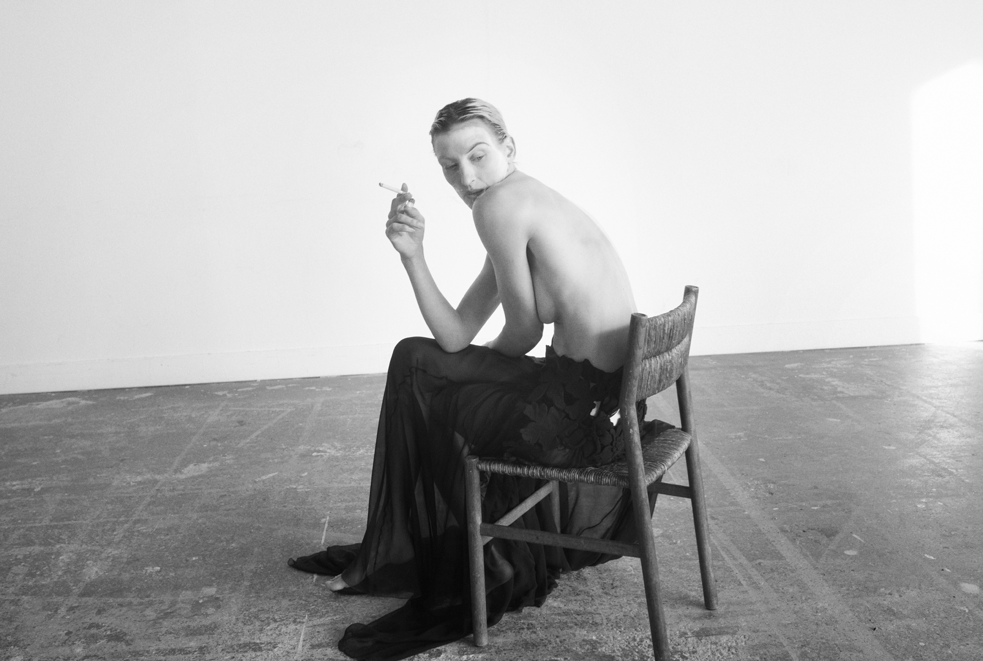 A woman is seated on a wooden chair topless. She is leaned over with a cigarette in hand with her chin pointed down and slightly covered by her shoulder. The model is photographed in an empty room. 