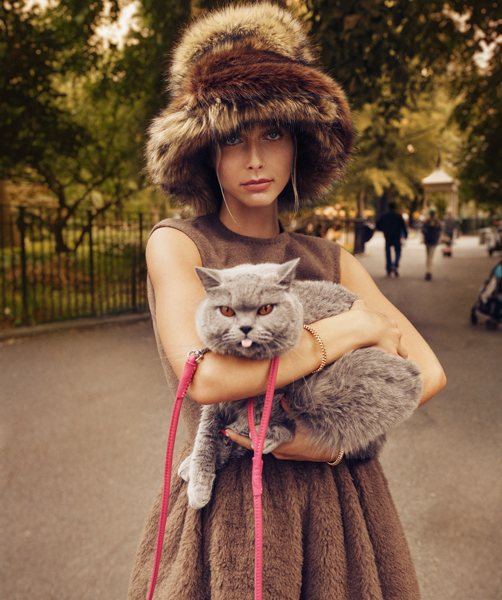 Emma Chamberlain is holding a fluffy grey cat with a leash. She is wearing a large fuzzy hat, and a soft brown dress. 