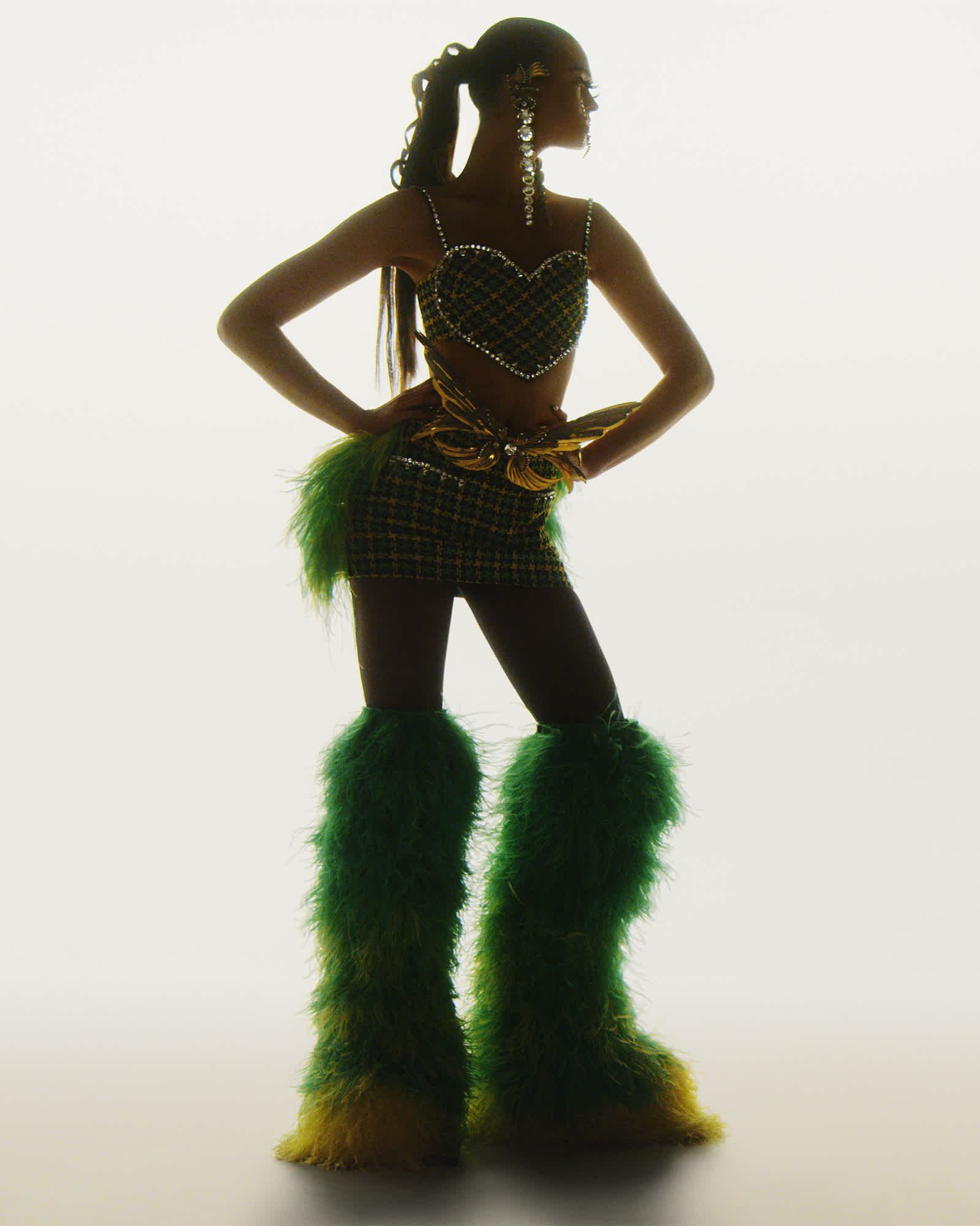 A woman is in the frame standing with her hands on her hips. She is in shadow partially with light illuminating from the background. She has her hair in a pony tail and is wearing furry knee high boots.