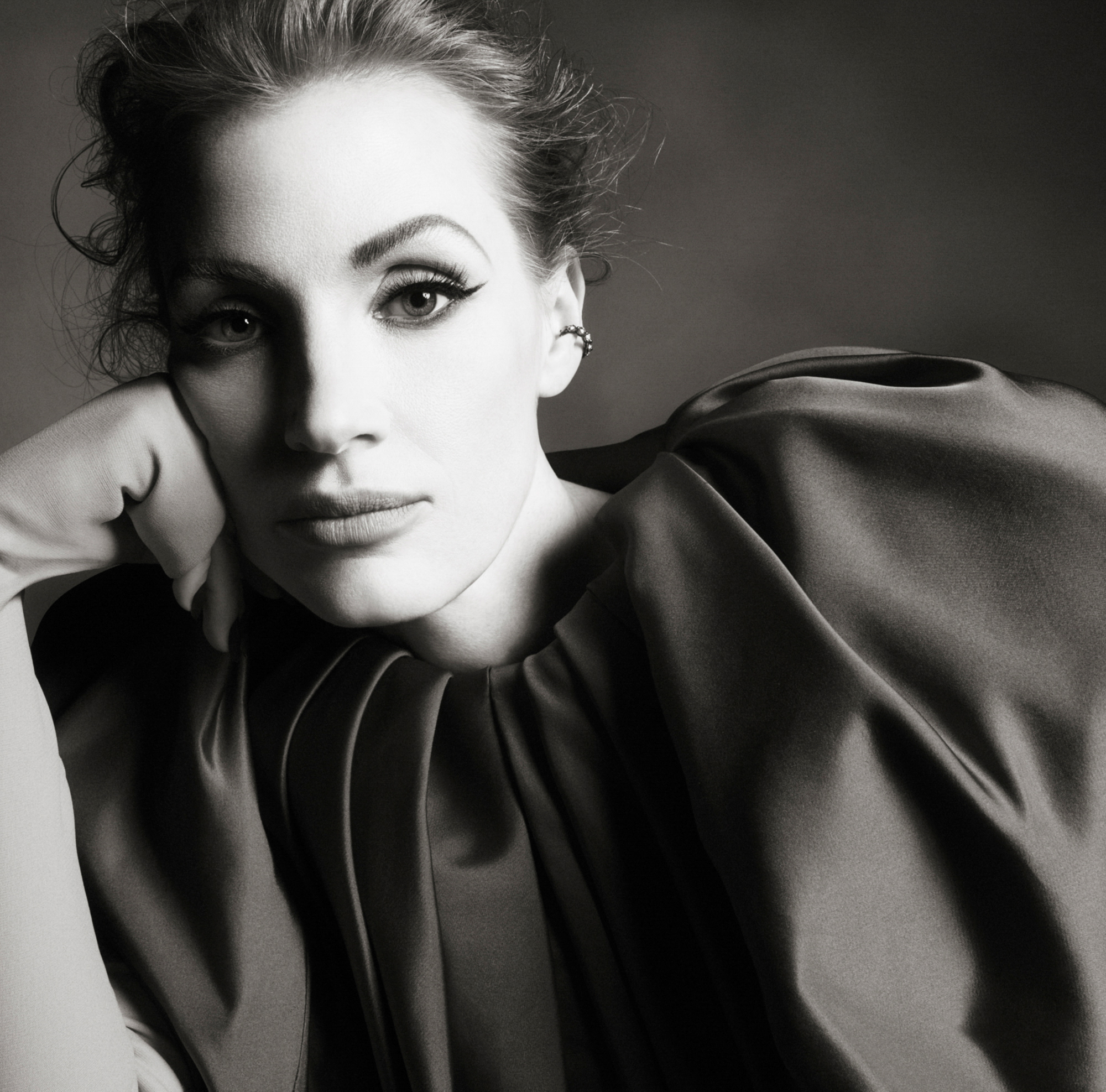 Jessica Chastain is wearing a dark silky garment and is leaning her head on her hand which is covered with fabric. She is looking directly at the camera, and her face is being contrasted with a shadow across half of her face. 