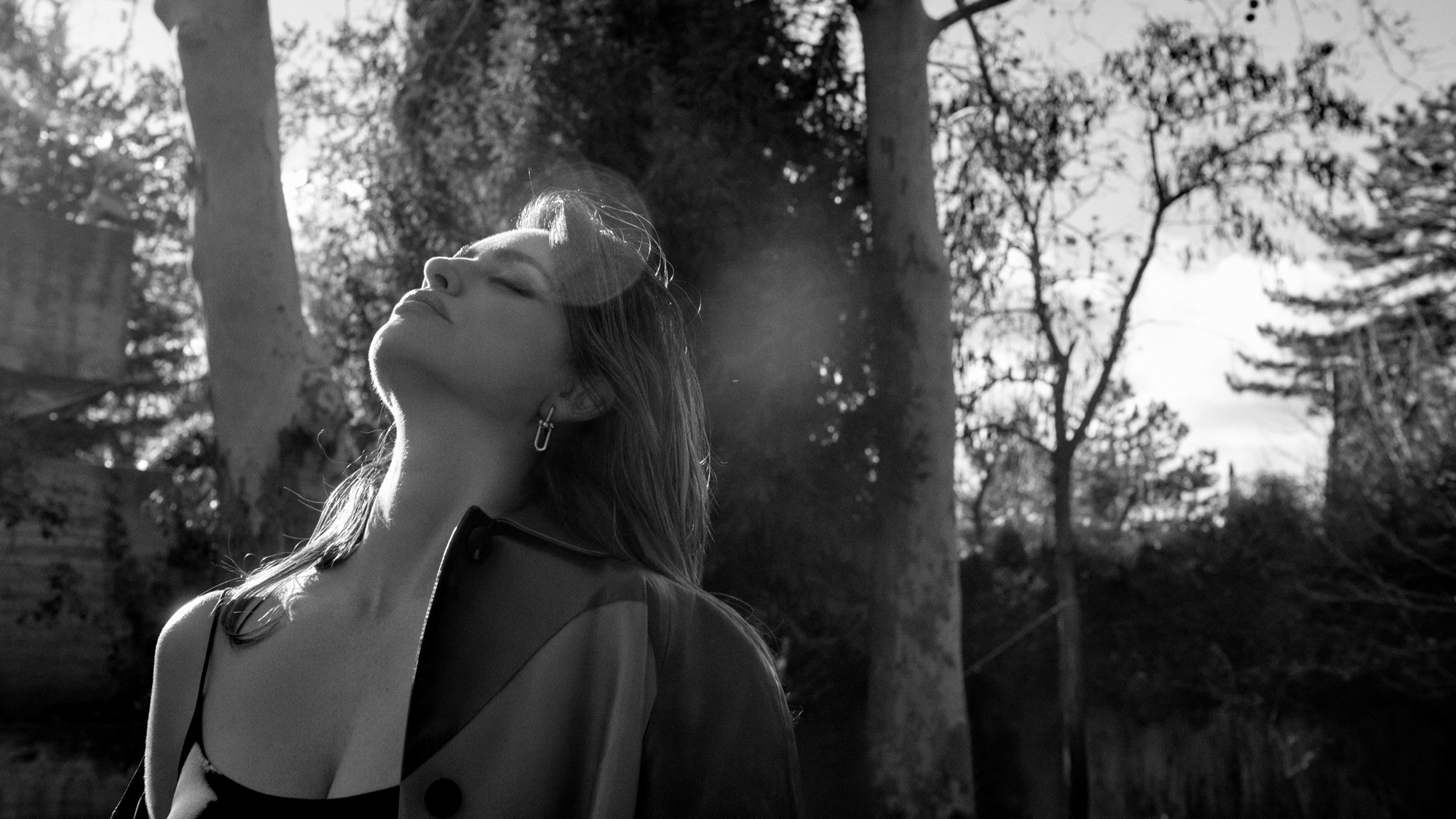 Penelope Cruz is standing outside with trees in the background. She is leaning her head back with her eyes closed, and has her jacket hanging on by one shoulder. 