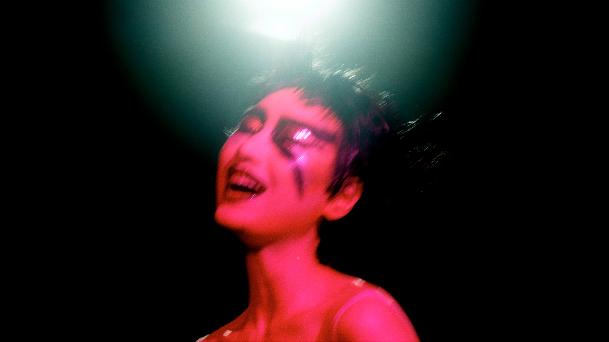 A woman is illuminated in pink toned light, she has angular dramatic eye makeup that extends down into the cheeks. There is shine on the mouth and eyelids, and her hair is in messy spikes. There is a spotlight shining more cool blue toned light over the model from the top of the frame. 