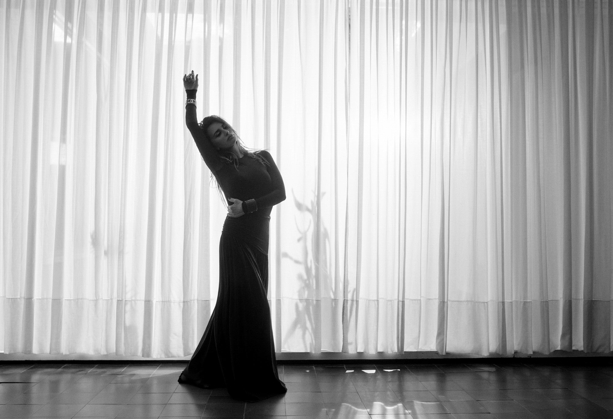 Penelope Cruz has her arm raised and is wearing a full length dress. She is standing in a room with square tiled floor and sheer curtains that are letting light through the windows. 