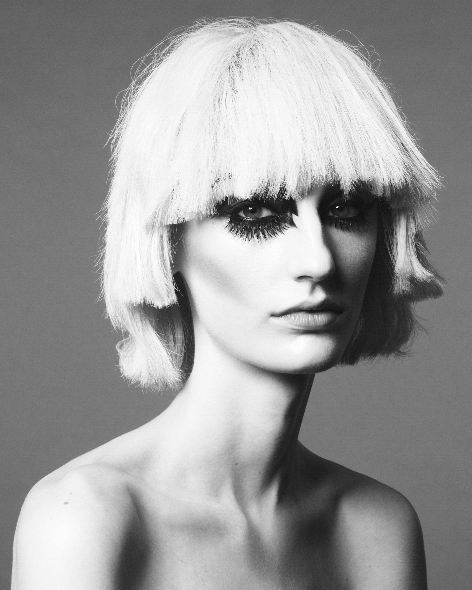 Model is featured in the center of the frame and is visible from the shoulders up. They have slight contouring, heavy false eyelashes, and a choppy more angular bob haircut. 