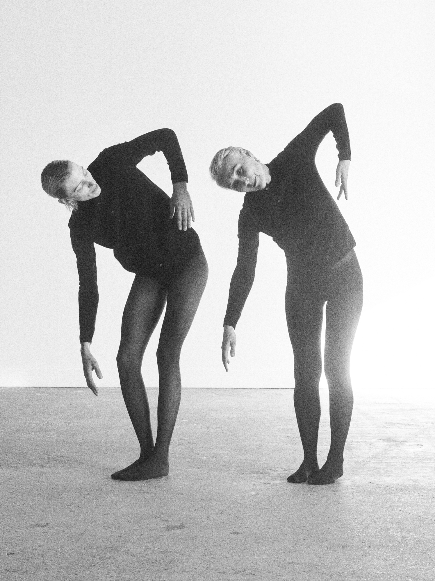 Two models are wearing fitted black garments. They both are leaning down and have their arms pointed downward. Light is flooding the blank room from the right hand corner. 