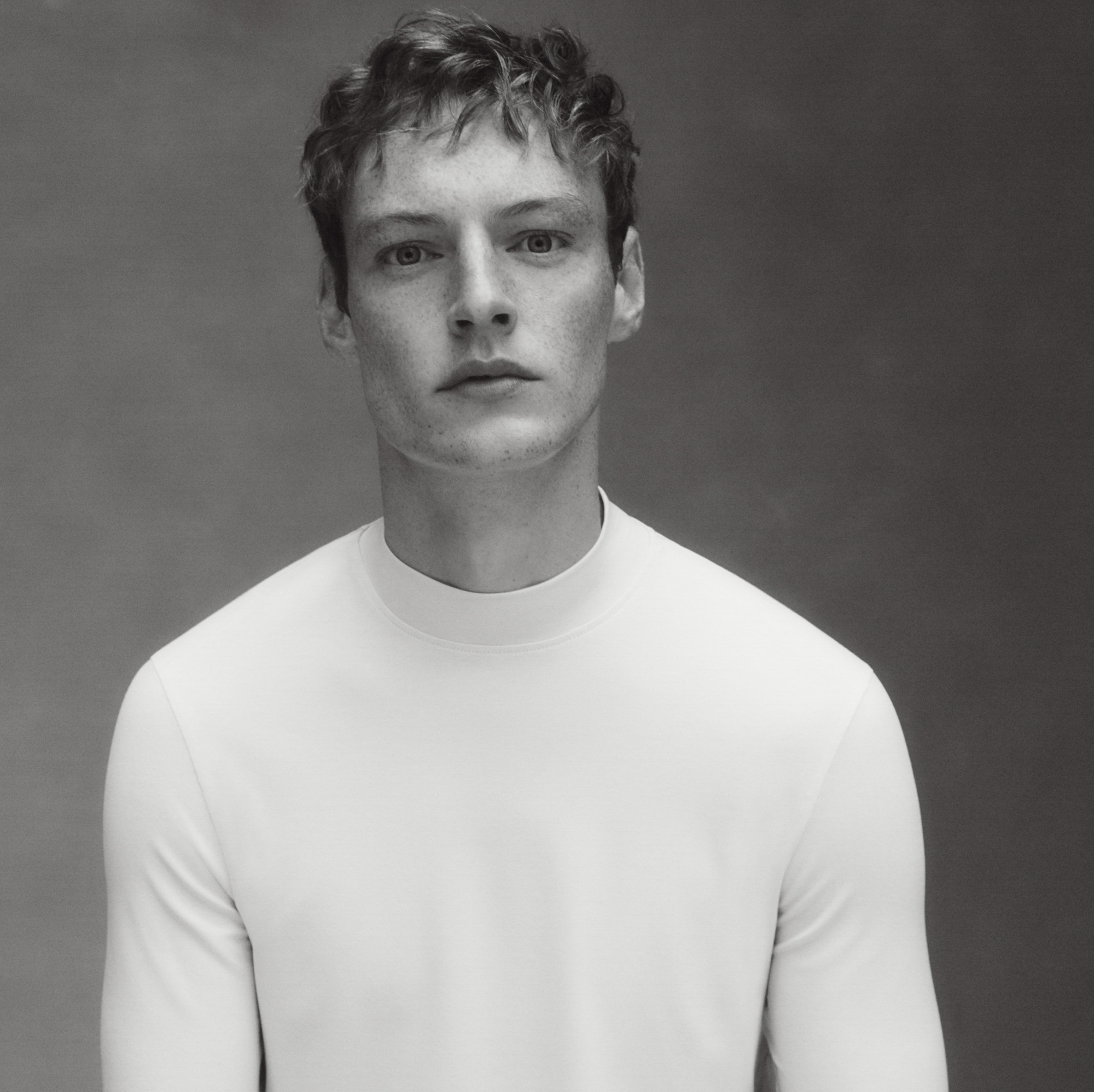 A man with reddish blonde hair is looking directly at the camera, his torso is slightly off center in the frame. He is wearing a white mock neck sweater and is in front of a textured gray background. 