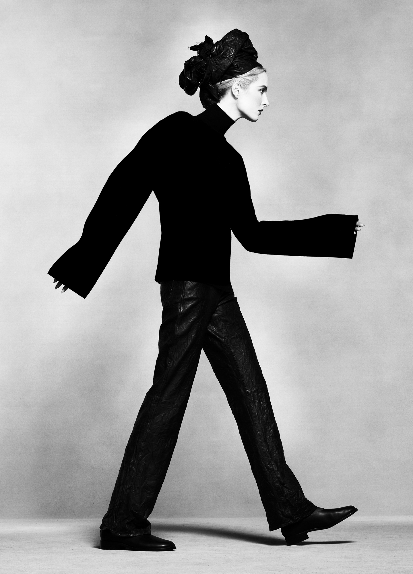 A model is posing in profile, she has her arms and leg extended as if she is walking forward. She is wearing all black, thick sweater, headpiece, pants, and boots. 