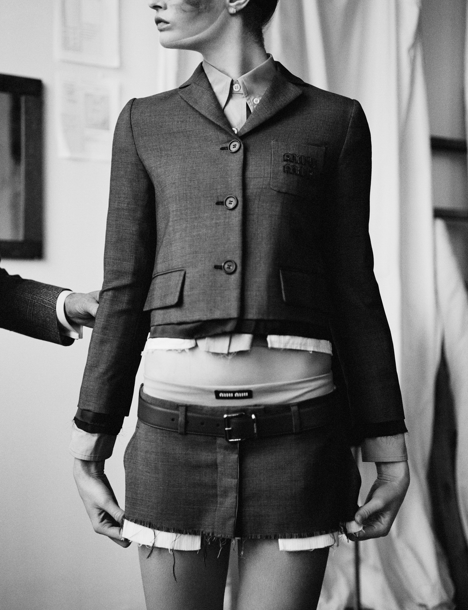 The model is standing in the center of the frame. She is wearing a Miu Miu jacket and skirt. An older man's hand is coming into the frame from the left hand side where he is touching a sleeve. The model's hands are resting at the bottom of the skirt. 