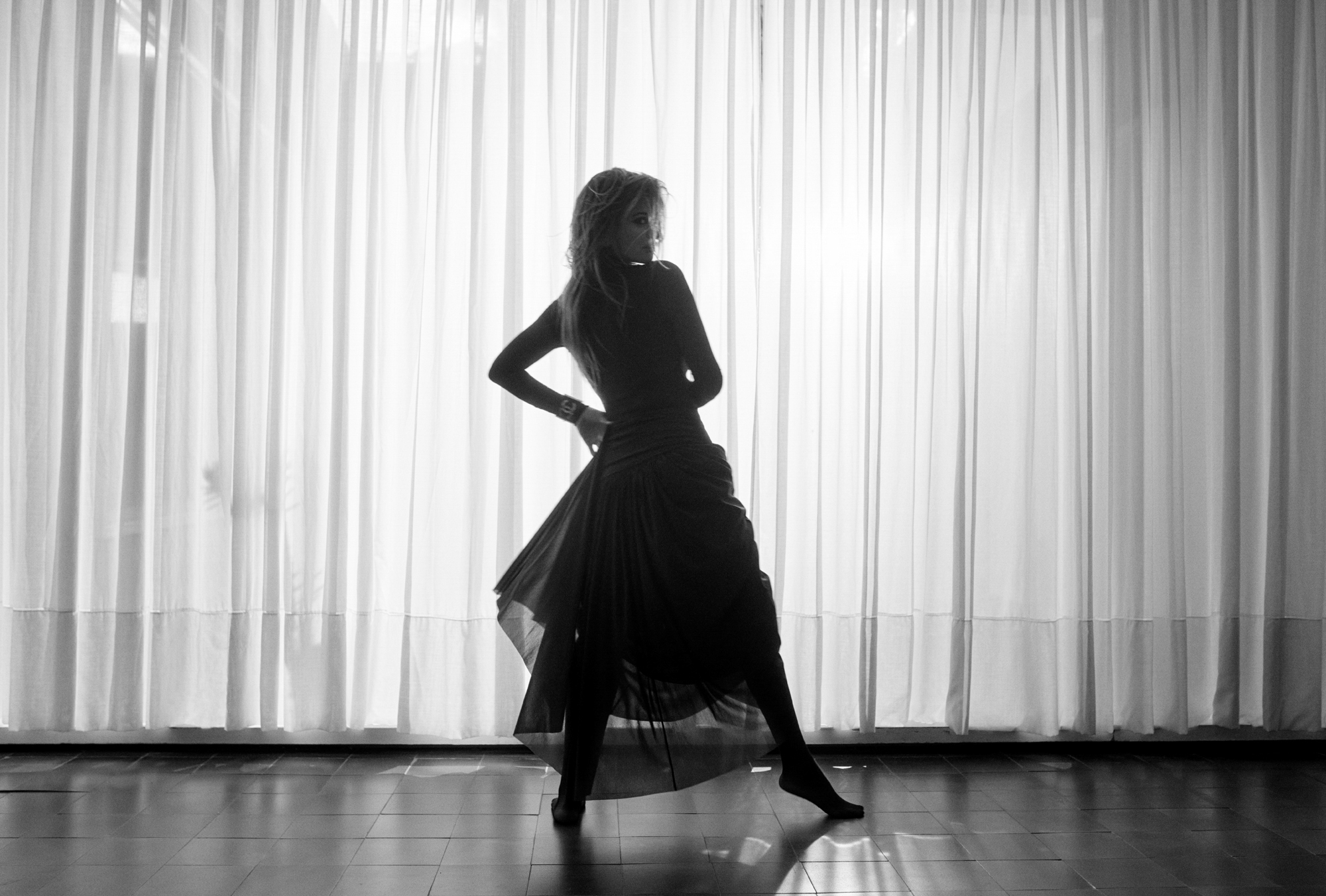 Penelope Cruz has her back turned away from the camera, and is looking backwards at the camera. She has one foot pointed as if in the middle of a dance. 