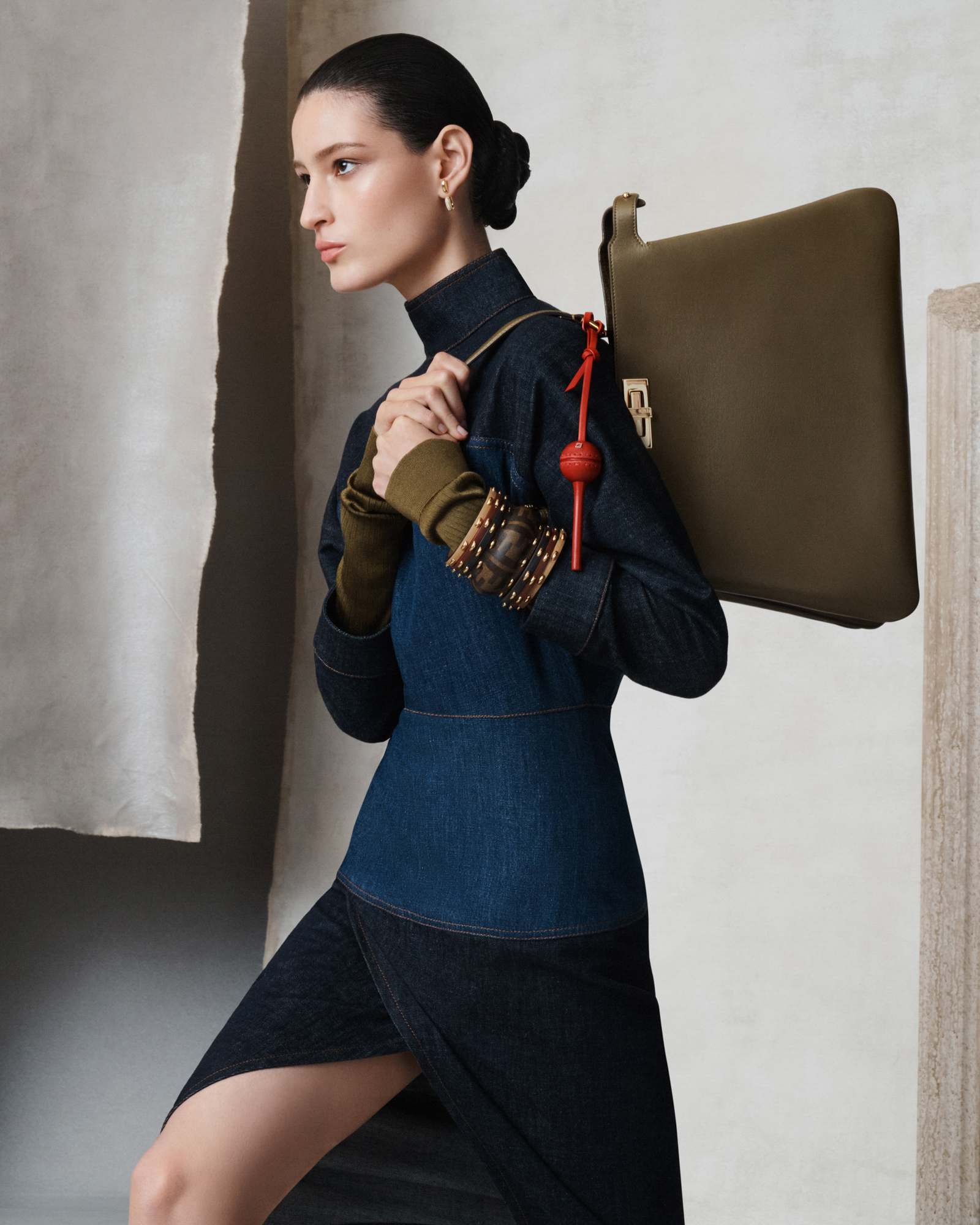 A woman is wearing a full length denim dress, and holding a medium sized dark green leather Fendi bag on her shoulder. Her hair is in a neat bun, and she has brown toned bracelets. The background is different kinds of grey paper, some textured, some smooth. A Greek column is visible on the right hand side of the image. 