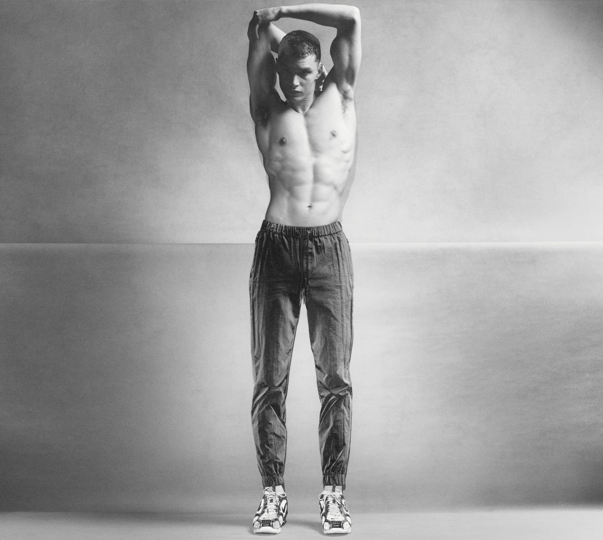 A man is standing in the center of the frame shirtless wearing textured athletic pants. He has on Nike running shoes. The background is split down the middle of the frame with one roll of grey background paper coming in front of the one behind it. The man is stretching his arms above his head. 