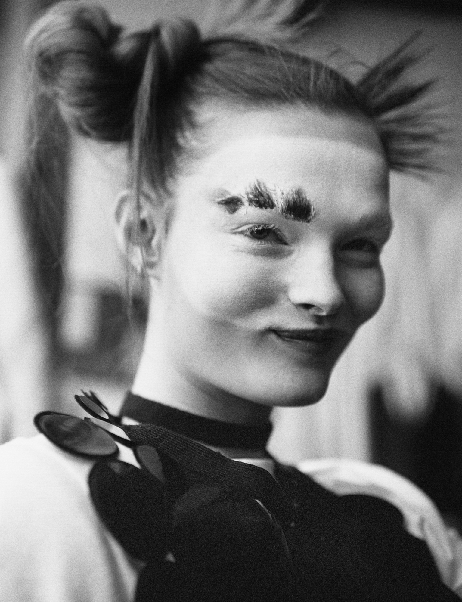 The image is a close up portrait of Lulu Tenney with mime like makeup up. Her hair is up in two top knot buns. She is smirking. 