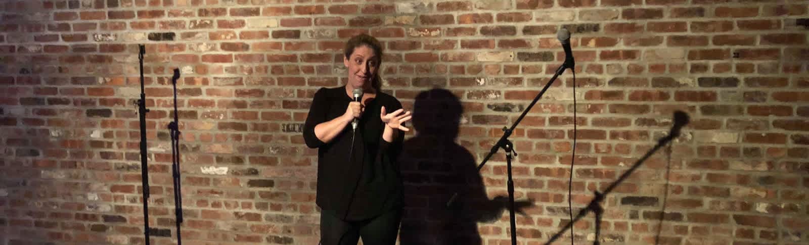 Pam standing in front of a brick wall with a mic doing stand up