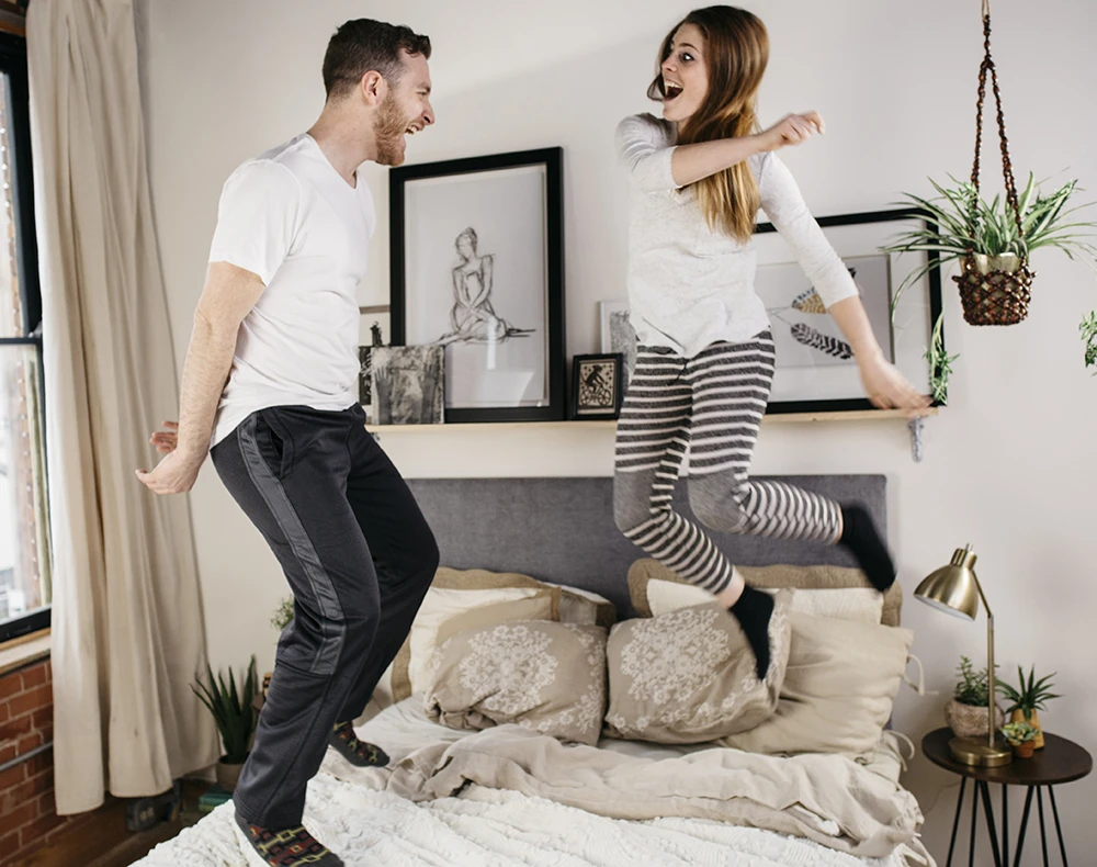 A young couple jumping on their white queen size metal Platform Bed with a blueish gray headboard in their urban apartment with exposed brick walls.