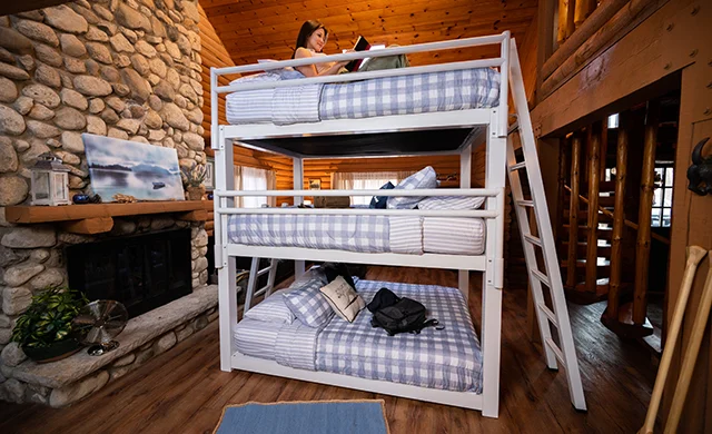 A young woman reading a book on the top bunk of a white queen size triple bunk bed for adults in a beautiful cabin.