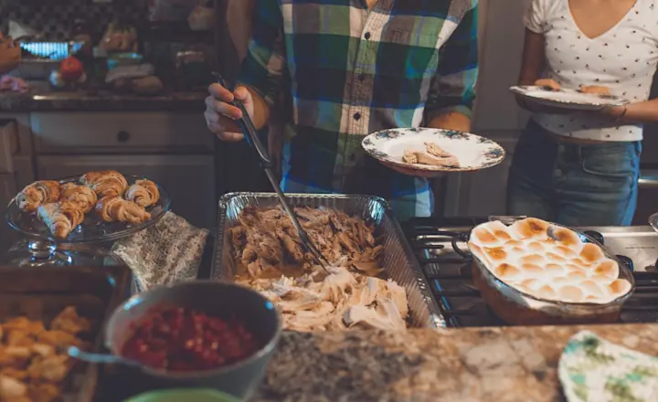 People serving food in a kitchen. Plates are filled with turkey, and there are side dishes like rolls, cranberries, and a marshmallow-topped sweet potato dish on the counter. The scene is casual and suggests a festive meal gathering.