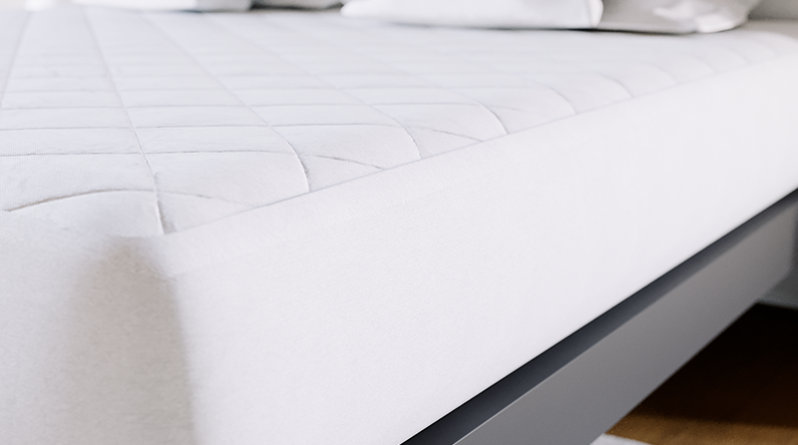 Super close up on a white bamboo mattress pad secured to a charcoal King size metal Platform Bed. Seen from the lower left-hand corner.
