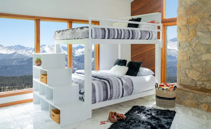 A modern bedroom with a white bunk bed and patterned bedding by large windows showing a mountain view. A stone wall is on the right, and a black rug is on the marble floor. Shelves with baskets and a cozy atmosphere complete the room.