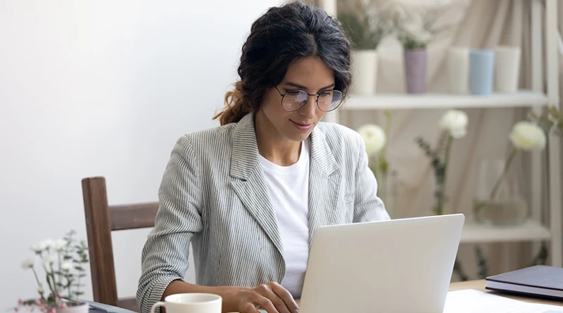 A millennial woman with dark curly hair in glasses and a light gray blazer working on a light gray laptop computer.