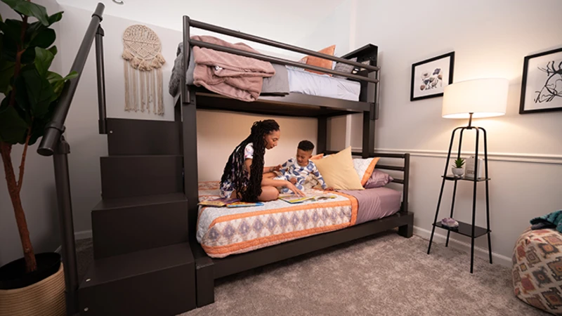 Young mother reading to her young son on the bottom bunk of a charcoal mixed size Adult Bunk Bed seen from a distance at the lower left hand corner of the bed frame.
