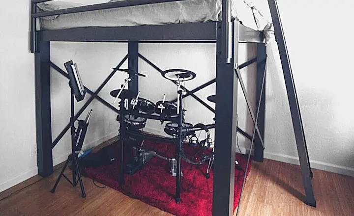 Lofted metal bed frame with a ladder in a minimal room. Underneath, there's an electronic drum set placed on a red rug. The floor is wooden, and the overall decor is simple and modern.