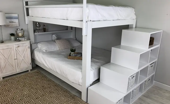 A modern, minimalist bedroom with a white loft bunk bed over a full bed. The stairs are cube shelves with woven baskets. A textured beige rug lies on the floor. Decor includes plants and coastal-themed artwork. Light walls enhance the airy feel.