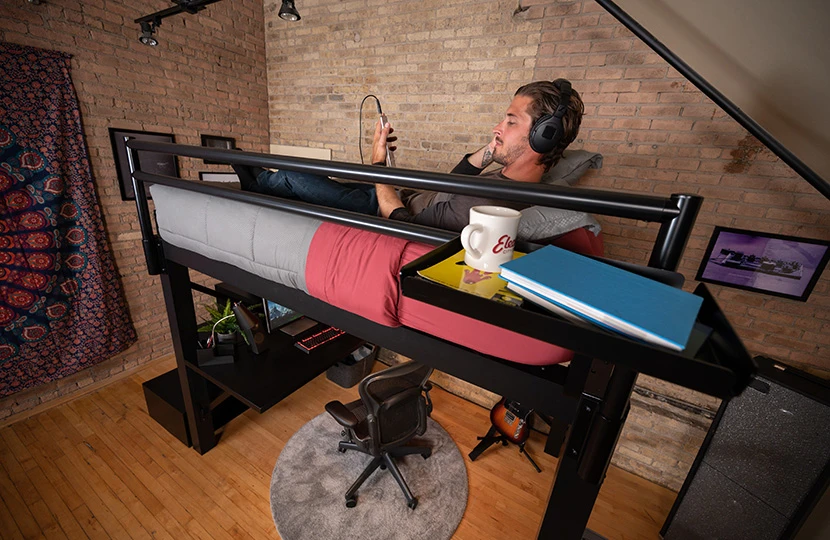 A man laying on a black Queen Adult Loft Bed resting his head on the pillows while listening to music on headphones and scrolling through his smartphone.