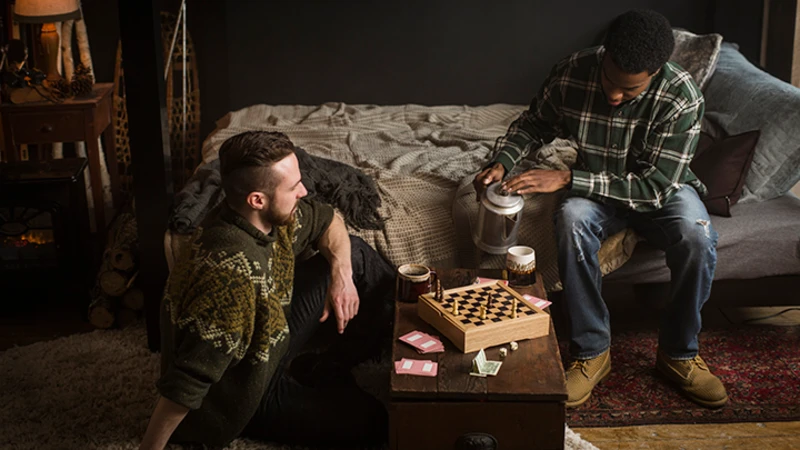 Two adult men in a ski lodge bedroom, with one sitting on the bottom bunk of a black Adult Bunk Bed pouring coffee into a mug on the table as his friend sits on the floor next to him.