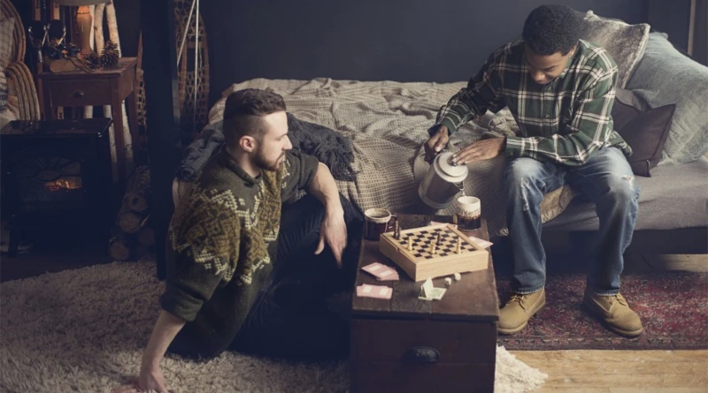 Two men drinking coffee together. One man sitting on the floor, the other on the bottom bunk of a black Queen Over Queen Adult Bunk Bed.