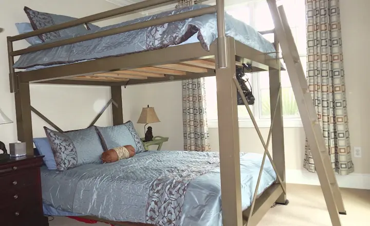 A bedroom featuring a loft bed with a double bed underneath. The bed has gray and blue bedding with decorative pillows. A small lamp and a bedside table are on the left, and a window with patterned curtains is in the background.