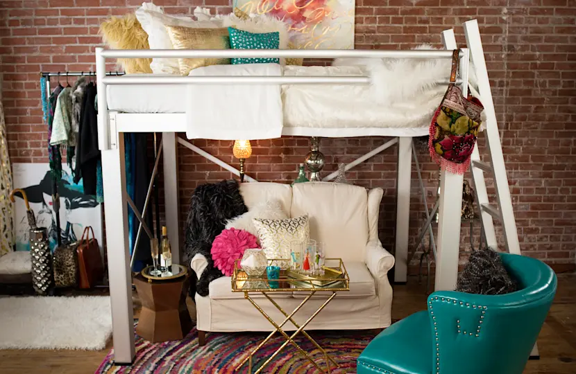 A stylish loft bed with fluffy pillows and a furry throw blanket overlooks a cozy seating area with a white armchair and a glass coffee table. A teal chair sits nearby on a colorful rug, and a clothing rack stands against a brick wall in the background.